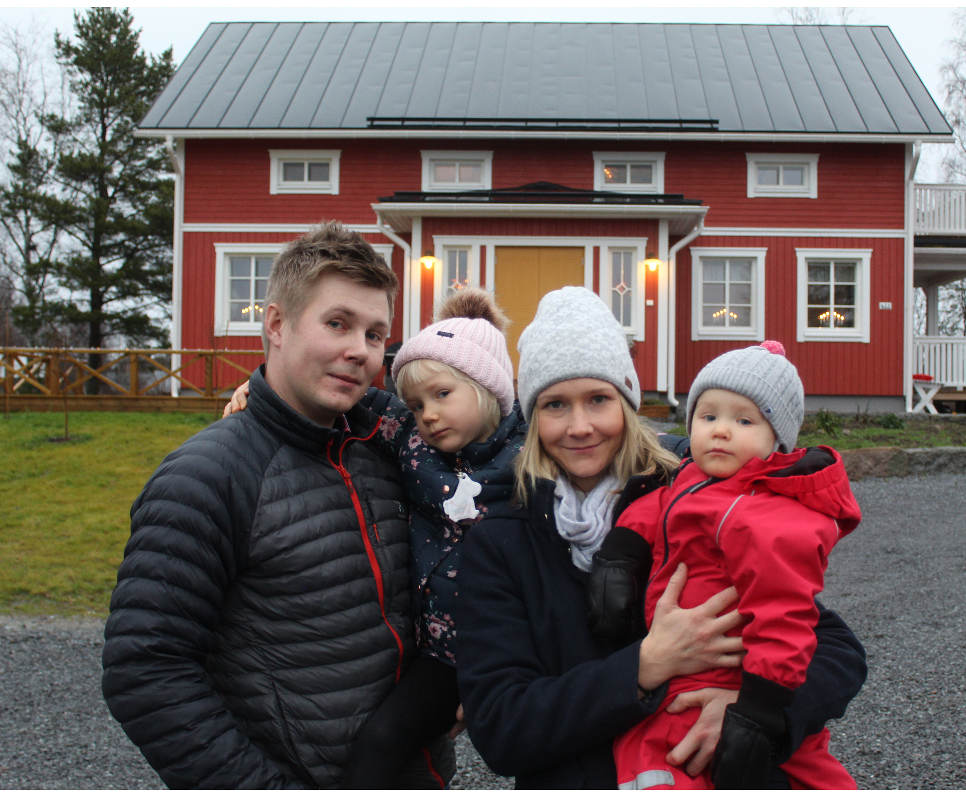 En familj med mamma, pappa och två döttrar står framför ett nybyggt rött hus i lantlig stil.