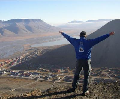 En man klädd i blå jacka med armarna utsträckta beundrar utsikten från ett berg.